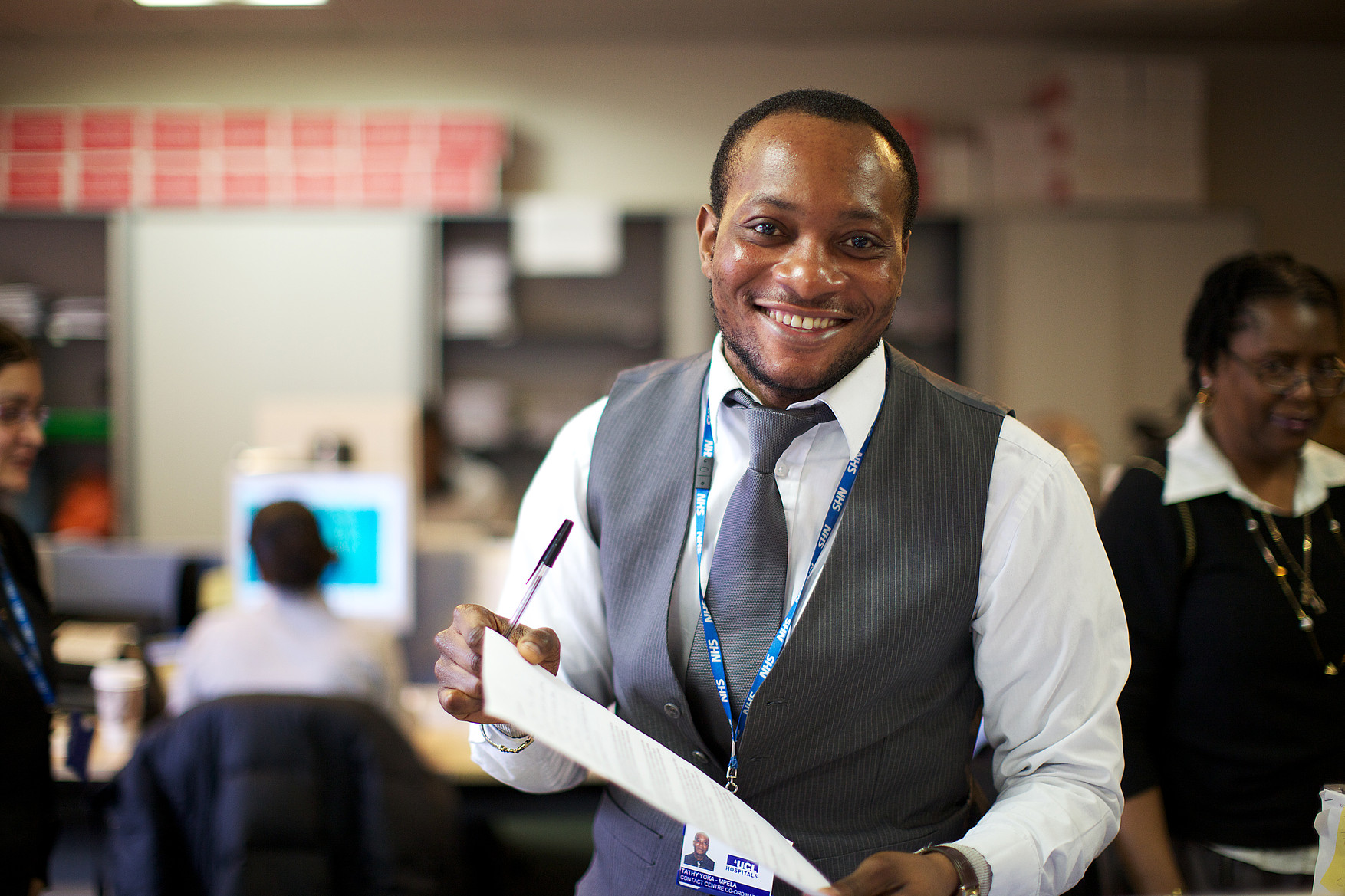 Unique ID: NHS_UCLH_DAY 1 CALL CENTRE_0045 Caption: Hospital administration, office, call centre. A member of staff looking at the camera, smiling, holding paperwork. People working in background. Restrictions: Copyright: ©