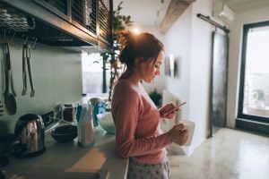 Young woman drinking first morning coffee and reading news in the kitchen of her apartment